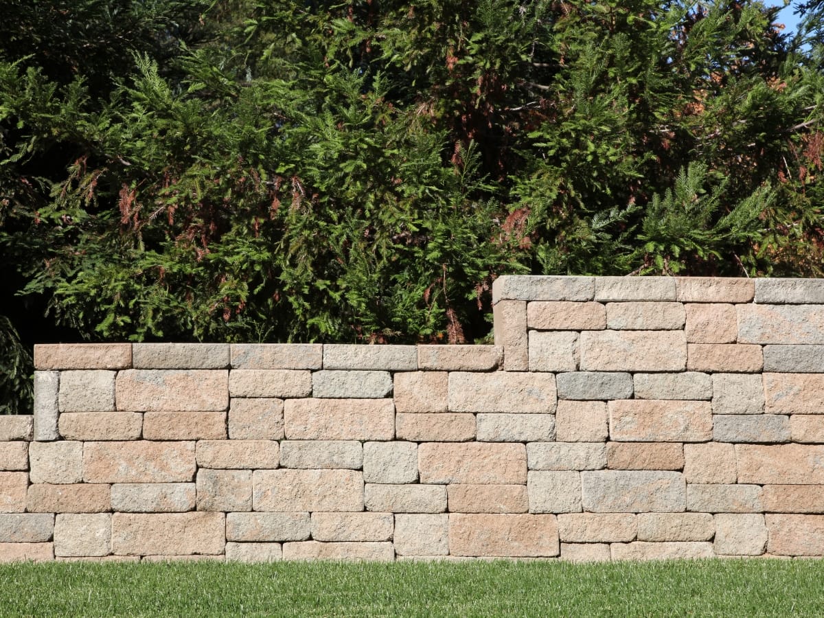How Commercial Retaining Walls in Sandy Springs Protect Your Business Property 15 A tan stone commercial retaining wall with an upper tier segment stands in front of dense green shrubs and a grass lawn.