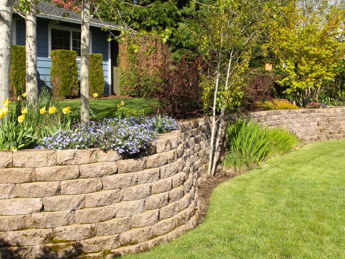 How Commercial Retaining Walls in Sandy Springs Protect Your Business Property 14 A neatly maintained yard with a stone retaining wall, flower beds, green grass, and trees in front of a house showcases the elegance often seen in commercial retaining walls.