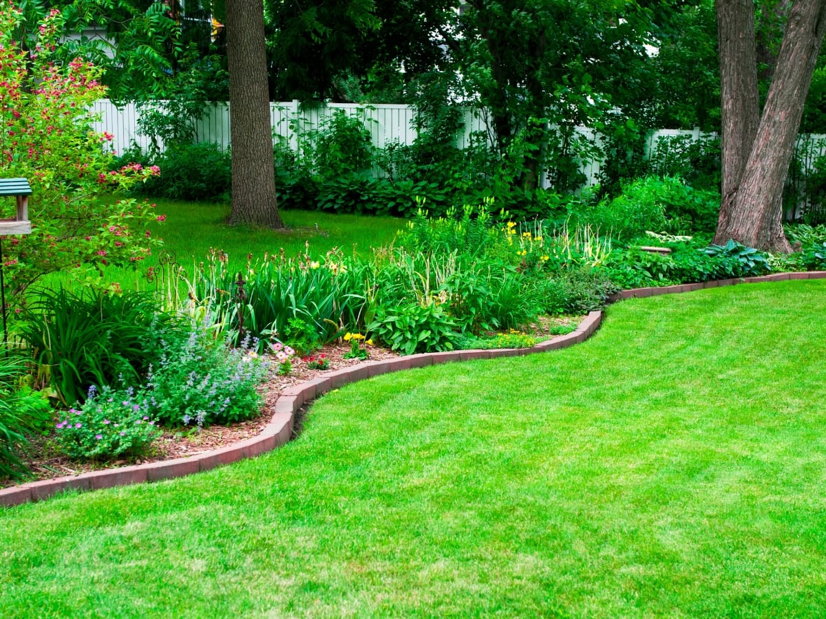 How Flo-Well Systems in Atlanta Can Improve Your Business Property’s Drainage 1 A well-maintained backyard garden with green grass, various plants and flowers along a curved mulched border, tall trees near a white fence, and discreet Flo-Well Systems in Atlanta for efficient drainage.