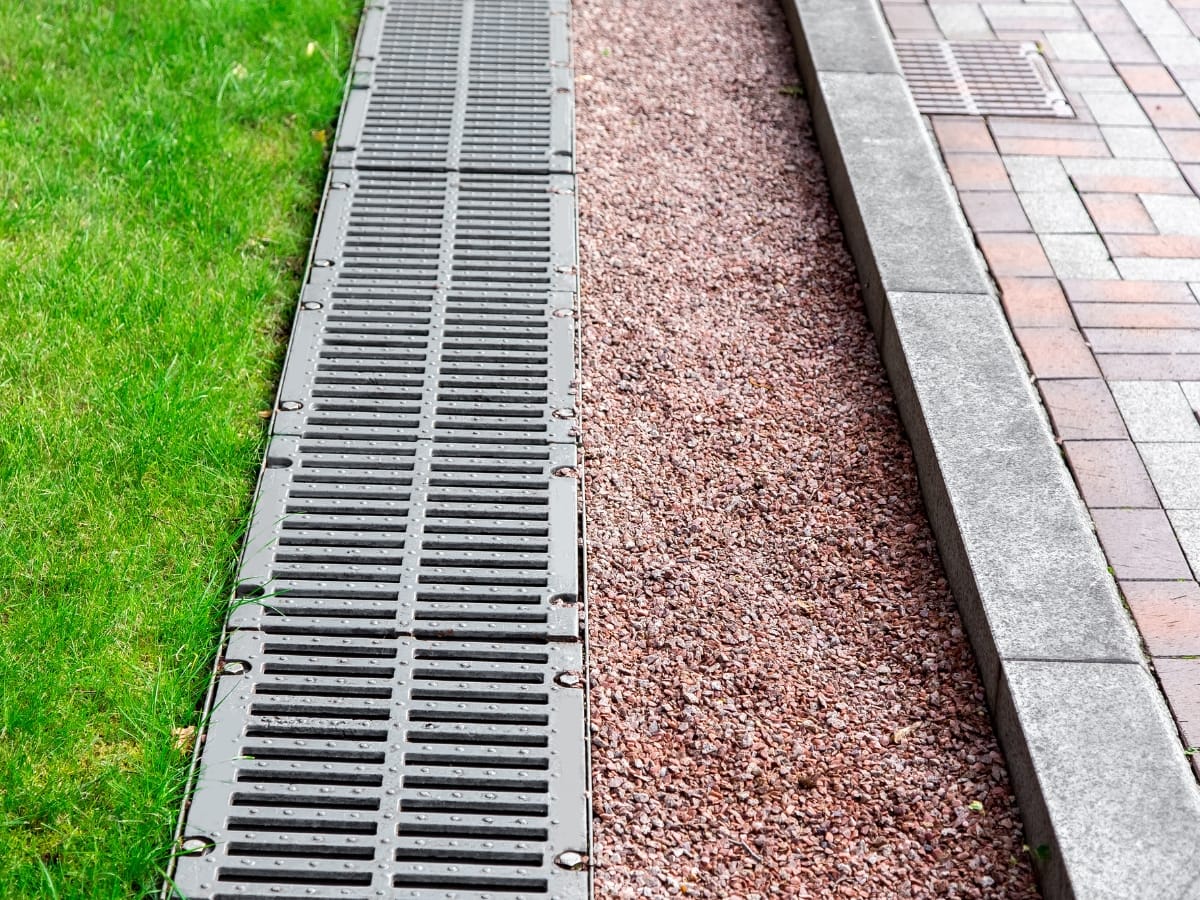 How Flo-Well Systems in Atlanta Can Improve Your Business Property’s Drainage 3 A metal drainage grate runs between a grass lawn and a gravel path, next to a brick sidewalk with a concrete curb, connecting seamlessly to Flo-Well Systems in Atlanta for effective water management.