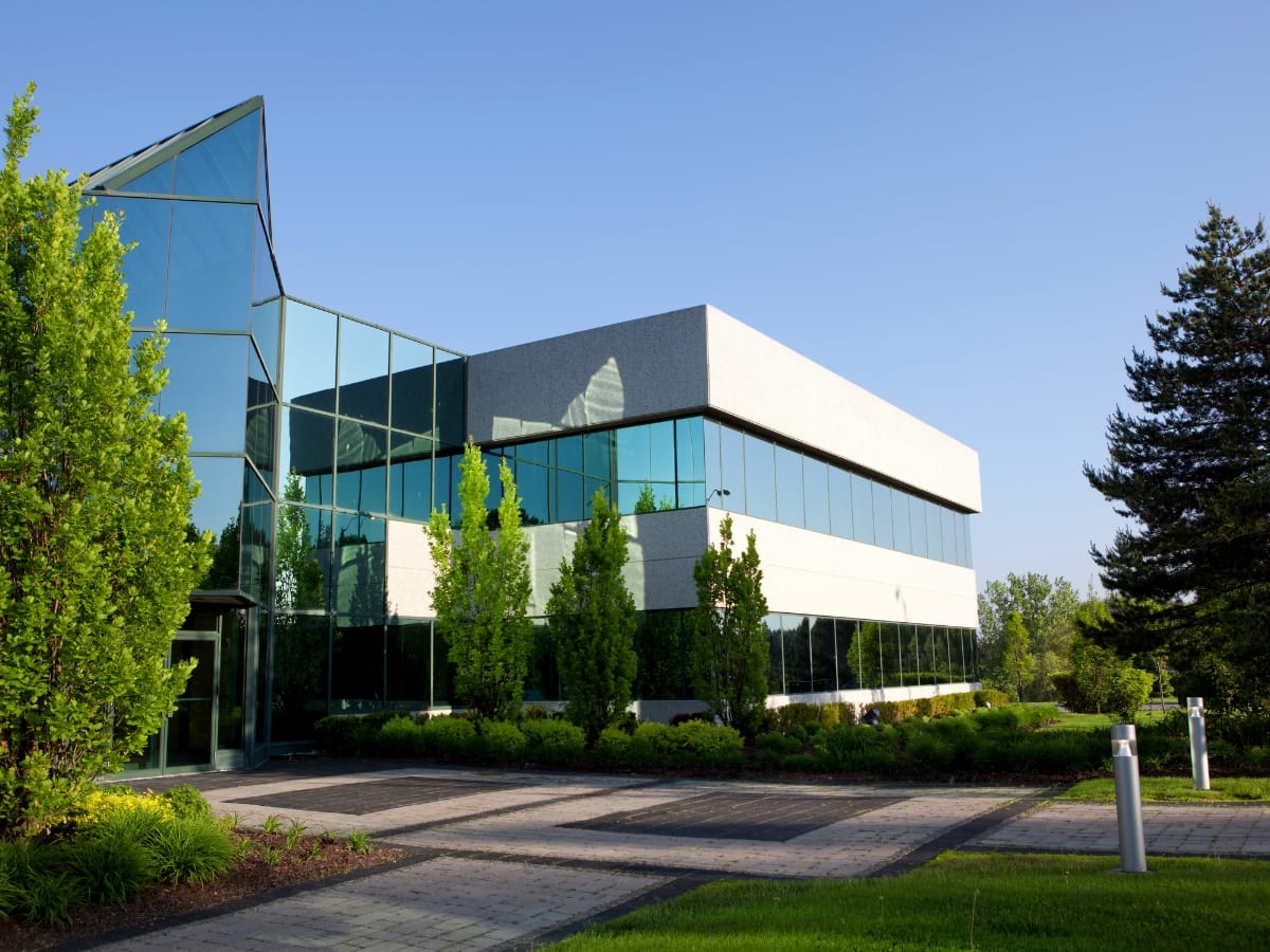 Enhancing Business Appeal with Hardscape Design in Woodstock GA 2 Modern office building with large glass windows, surrounded by green trees, landscaped lawns, and expert hardscape design in Woodstock GA, all beneath a clear blue sky.
