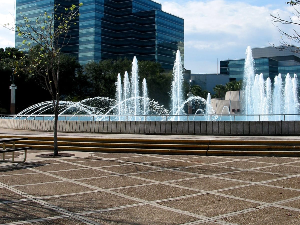Enhancing Business Appeal with Hardscape Design in Woodstock GA 4 A modern fountain with multiple water jets is surrounded by concrete benches and trees, showcasing expert hardscape design in Woodstock GA, with sleek glass office buildings visible in the background.
