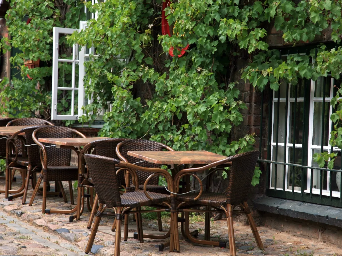 Enhancing Business Appeal with Hardscape Design in Woodstock GA 9 Outdoor café tables and chairs are set on a cobblestone patio, showcasing expert Hardscape Design in Woodstock GA, next to a building covered with green vines and open windows.