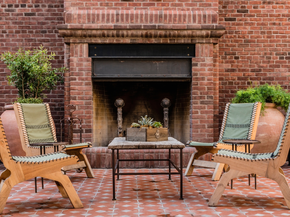 How Outdoor Fire Features in Atlanta Make Business Spaces More Inviting 3 Four wooden chairs and a small table sit on a tiled patio in front of a brick fireplace, showcasing stylish outdoor fire features in Atlanta, with large potted plants on either side.