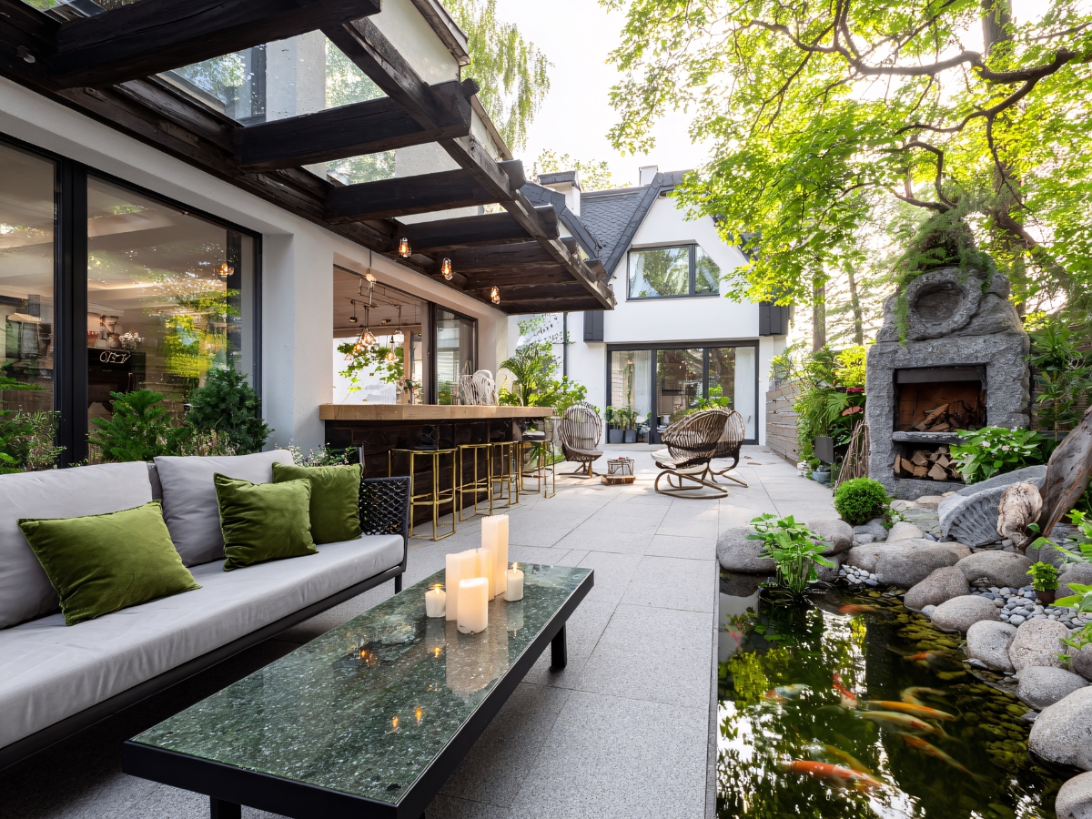How Outdoor Fire Features in Atlanta Make Business Spaces More Inviting 1 Modern outdoor patio with a sofa, glass table, bar area, stone fireplace—showcasing Outdoor Fire Features in Atlanta—a koi pond, and lush greenery beside a contemporary house.