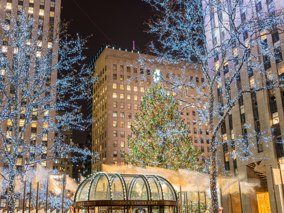 Holiday Landscaping Services Roswell GA Businesses Need 3 A large Christmas tree decorated with lights stands in front of tall buildings at night, surrounded by trees wrapped in string lights—an inspiring scene for those seeking Holiday Landscaping Services Roswell. The entrance to Rock Center Cafe is visible nearby.