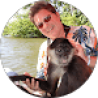 A person wearing sunglasses and a floral shirt is holding a monkey while sitting in a boat on a body of water, creating the perfect scene for an exotic vacation. A backdrop of trees completes this picturesque moment, reminiscent of scenes from travel testimonials.